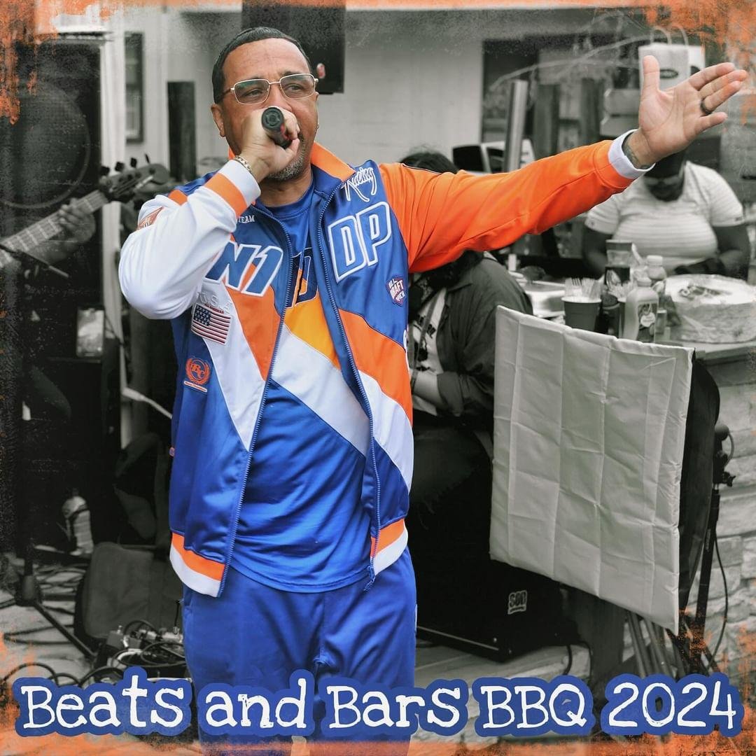 An MC in an orange-and-blue jacket reaches out with the mic, 2024.