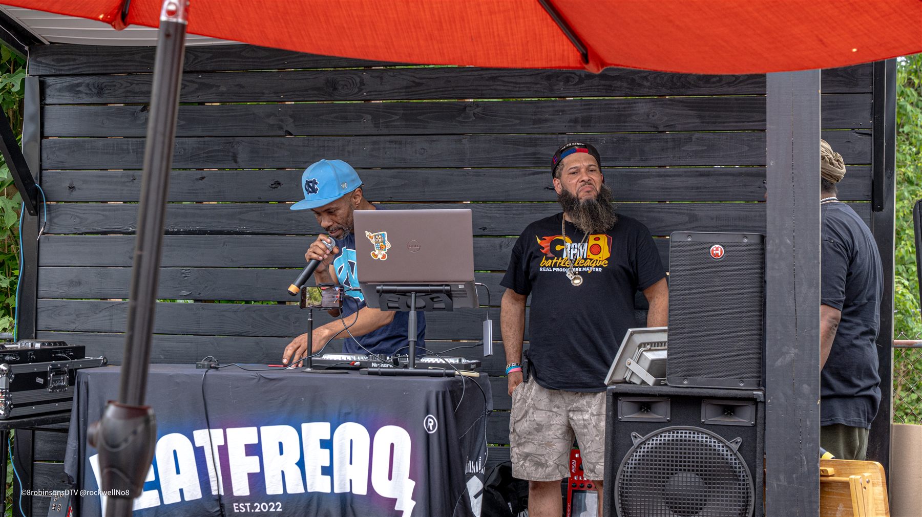 Two men at the Beat Freaq table under a red umbrella: one raps into a microphone over a laptop and DJ controller while the other stands beside him, watching.