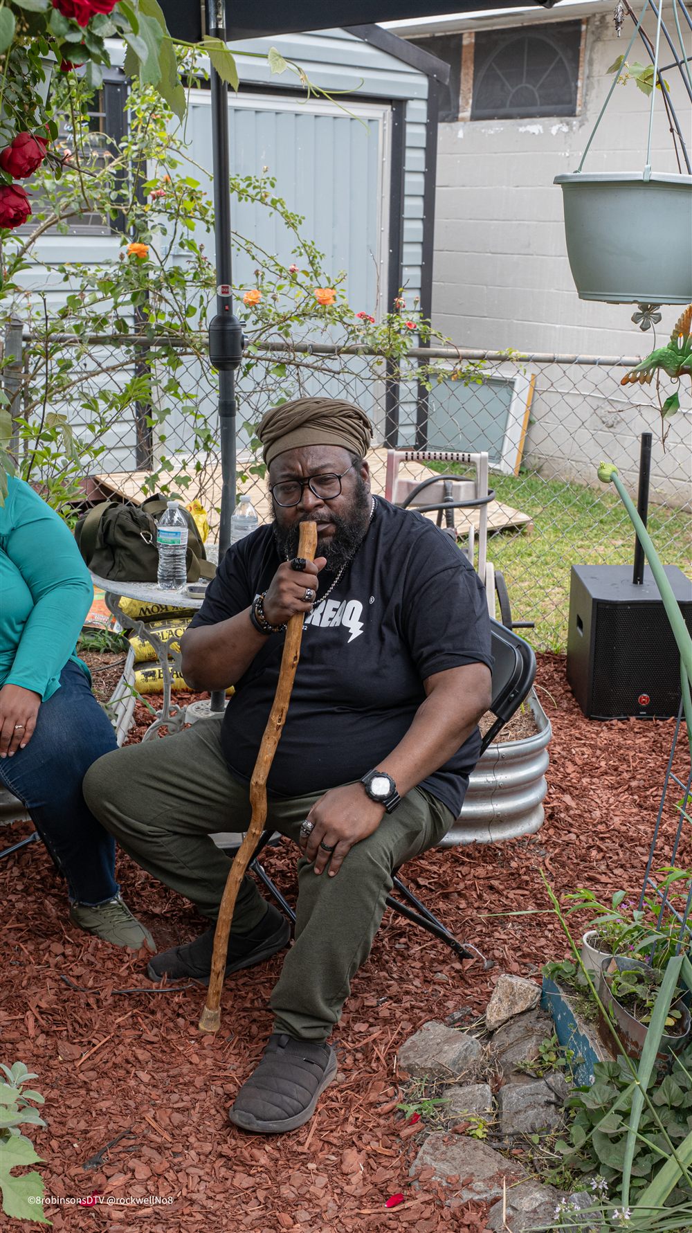 Jah Freedom, in glasses and an olive head wrap, sits in a folding chair in a garden with a wooden walking staff raised to his mouth.