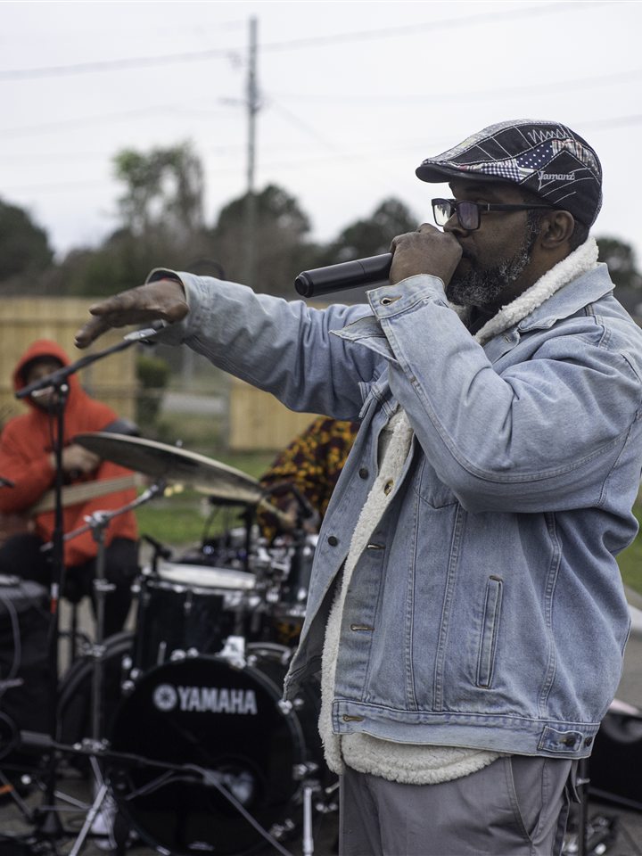 An MC in a denim jacket and flat cap on the mic with the band behind him, 2023.