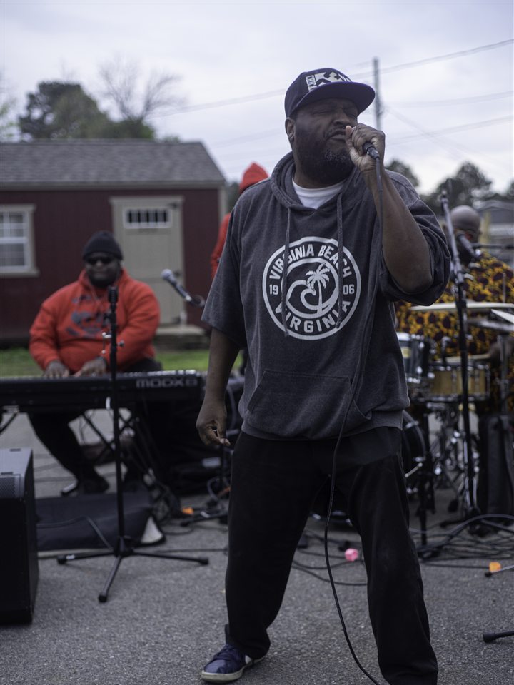 An MC in a gray hoodie on the mic, with the band behind him, 2023.