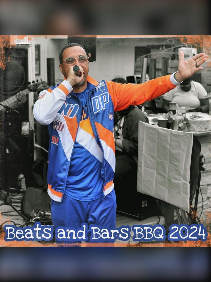 An MC in an orange-and-blue jacket reaches out with the mic, 2024.
