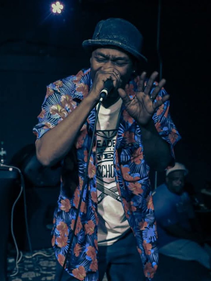An MC in a bucket hat and floral shirt on the mic, 2025.