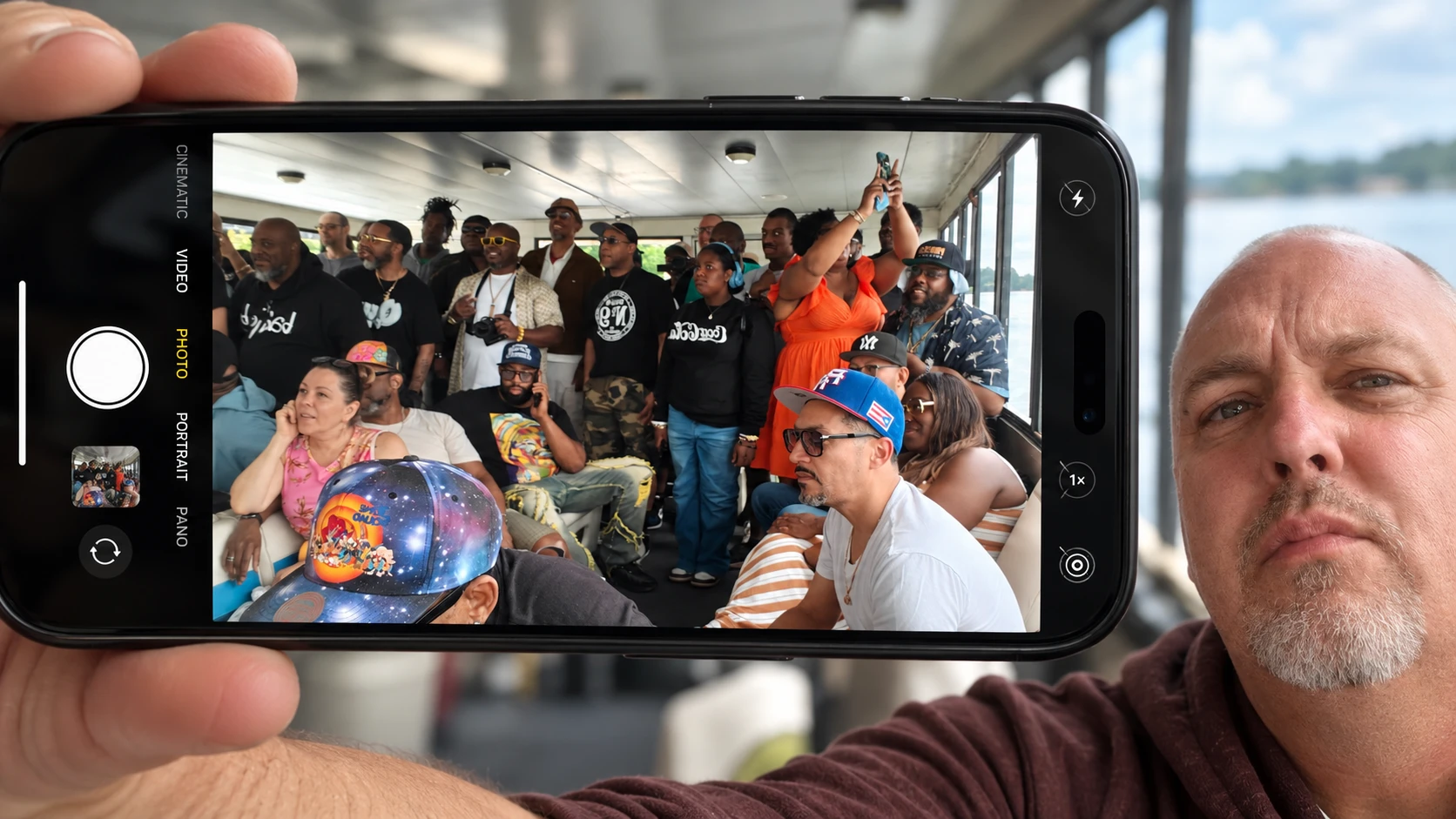 A man holds up a phone in landscape. Its camera screen shows a crowd of people gathered inside a boat cabin, with the water visible through the windows behind him.